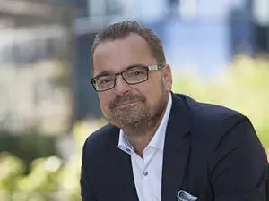 Natural Pharmaceuticals Melatonina Vidutinio amžiaus vyras su akiniais ir kostiumu prie neryškaus miesto fono.