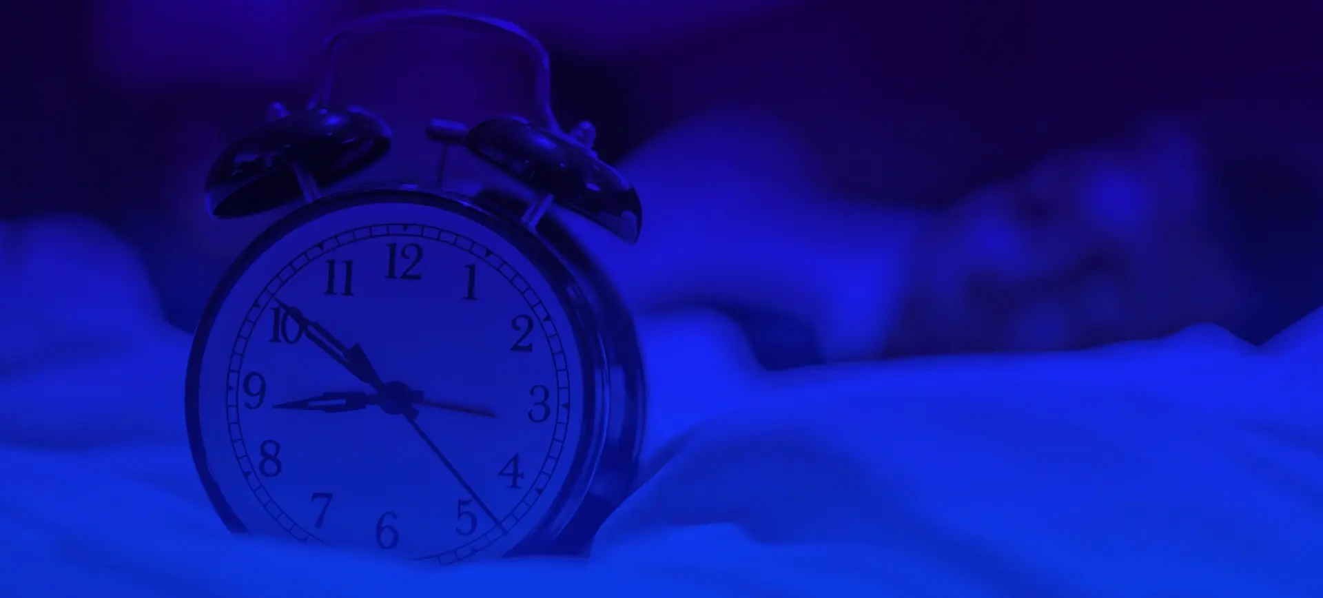 Natural Pharmaceuticals Melatonina Nuotraukoje matomas klasikinis žadintuvas su varpeliais. Fone, neryškiai ir vos matomas, guli lovoje miegantis žmogus. Visą sceną apšviečia intensyvi mėlynai violetinė šviesa, kuri suteikia jai naktinę ir ramią atmosferą.