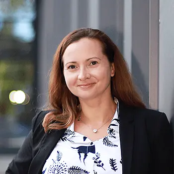 Natural Pharmaceuticals Melatonina Vidutinio amžiaus moteris dėvi juodą švarką ir baltą palaidinę su augaliniais raštais, šypsosi ir stovi prie sienos.